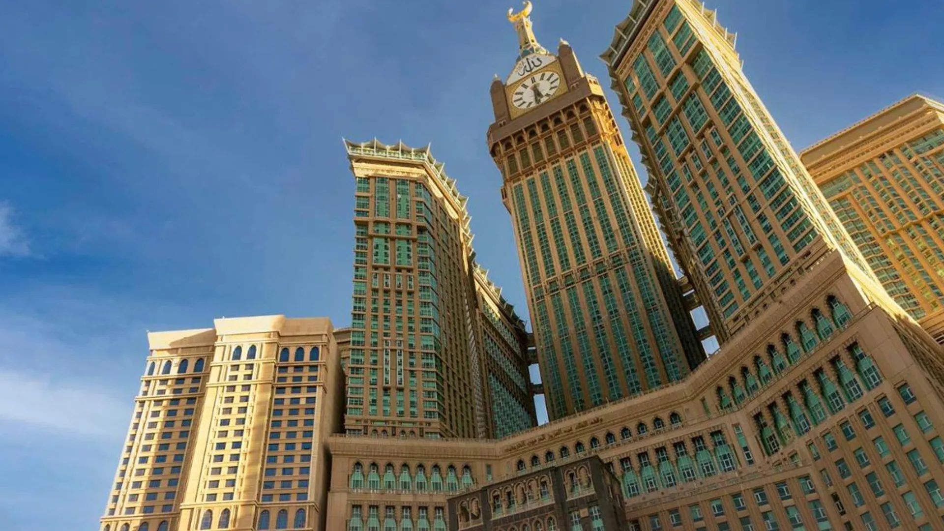 أبراج شاهقة ذهبية اللون لمجمع أبراج البيت في مكة المكرمة بالمملكة العربية السعودية، يهيمن عليها برج الساعة المركزي المتوج بهلال. تتميز المباني بمزيج من العناصر المعمارية الحديثة والإسلامية، مع نوافذ ذات لون أخضر وتفاصيل مزخرفة. يقف المجمع أمام سماء زرقاء صافية، مُظهراً حجمه الهائل وعظمته كمعلم بارز في المدينة المقدسة.