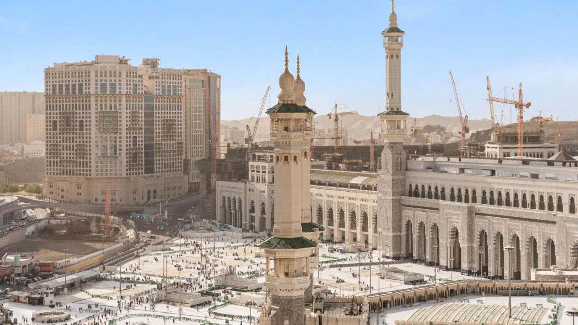 منظر جوي للحرم المكي الشريف والمنطقة المحيطة به في مكة المكرمة. يظهر في الصورة المسجد الحرام بأروقته وساحاته الواسعة المليئة بالمصلين، وتحيط به المباني الحديثة والأبراج الشاهقة، بما فيها برج الساعة الشهير. تظهر أيضاً أعمال التوسعة والبناء الجارية حول الحرم، مع وجود الرافعات ومواقع البناء. المشهد يجمع بين العمارة الإسلامية التقليدية والتطور العمراني الحديث، ويعكس أهمية المكان المقدس وحجم المشروعات التطويرية فيه.